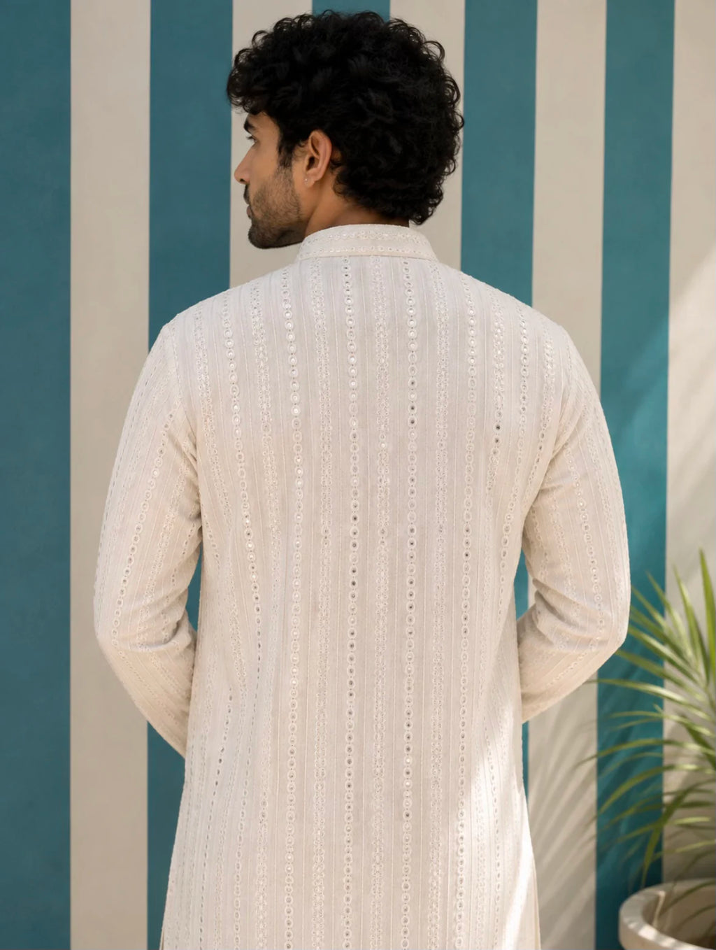 A man viewed from behind wearing a cream-colored, long-sleeved textured kurta with vertical eyelet embroidery, set against a blue and white striped wall.