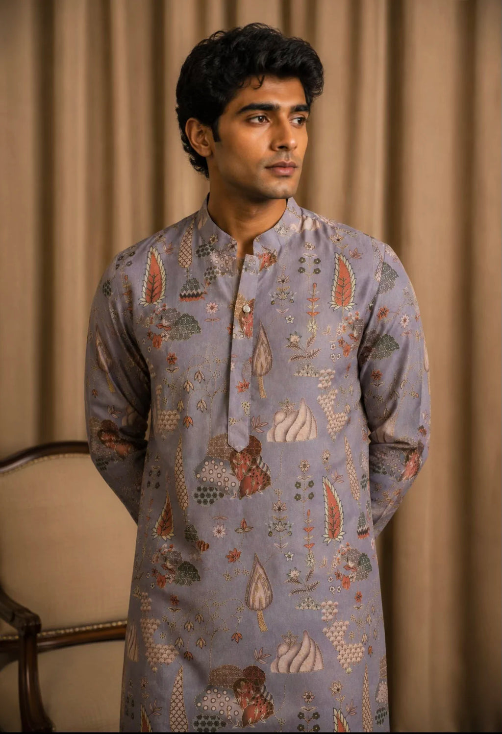 A man wearing a light blue-gray patterned kurta with traditional motifs, standing against a beige curtain backdrop.