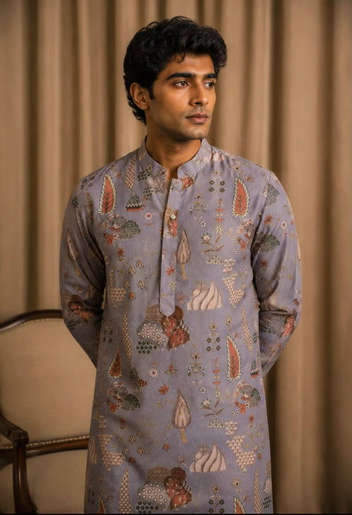 A man wearing a light blue-gray patterned kurta with traditional motifs, standing against a beige curtain backdrop.