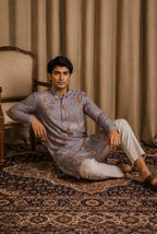 A man wearing a light blue patterned kurta and white trousers sits on a patterned rug in front of beige curtains, with a wooden armchair visible on the left.