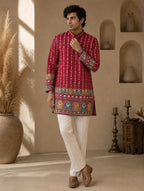A man modeling a red long-sleeve kurta with intricate multicolor embroidery and vertical white stripes, paired with white trousers and brown shoes, in a warm, rustic interior setting.