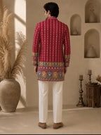 A man viewed from behind wearing a bright red kurta with vertical white stripes and a colorful embroidered border, paired with white trousers, in a warm beige interior setting.