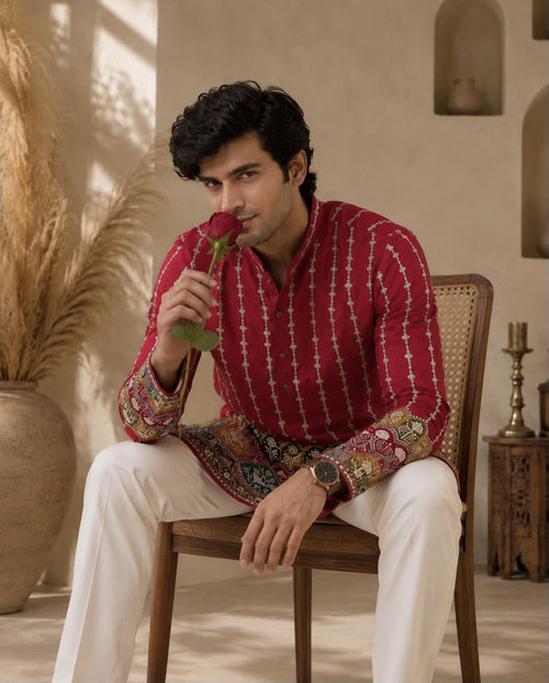 A man sits on a wooden chair wearing a red and gold patterned kurta with white trousers, holding a red rose near his face in a warm, styled interior setting.