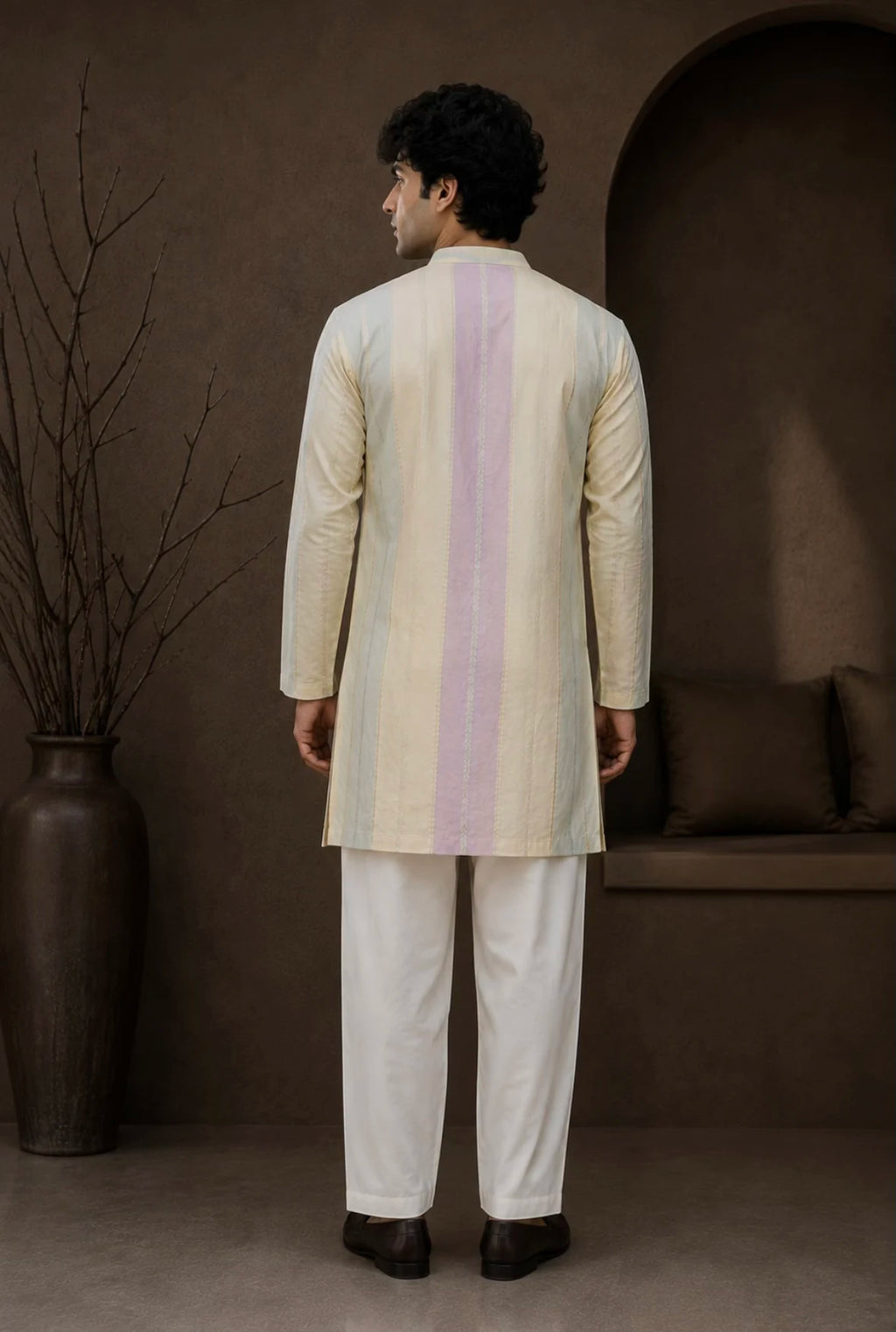 A man viewed from the back wearing a cream-colored kurta with a vertical pastel stripe in light purple and beige, paired with white trousers, standing in a styled studio setting with a brown background and a large vase.