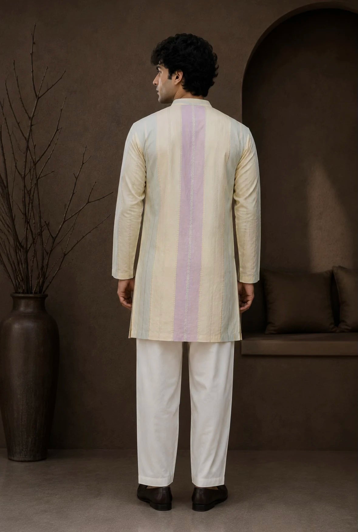 A man viewed from the back wearing a cream-colored kurta with a vertical pastel stripe in light purple and beige, paired with white trousers, standing in a styled studio setting with a brown background and a large vase.
