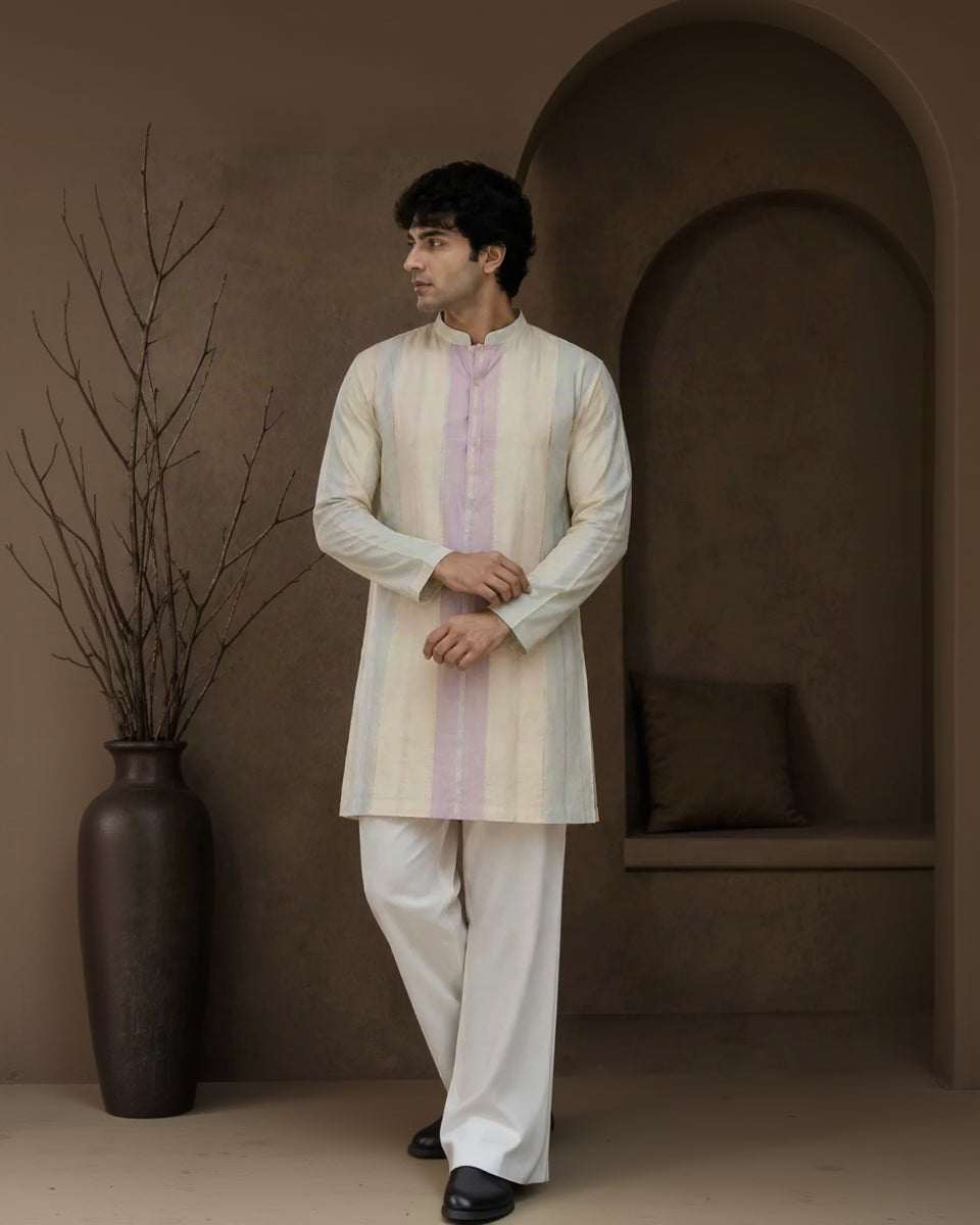 A man modeling a traditional Indian outfit consisting of a cream and light purple striped kurta with white pants, standing in a rustic indoor setting with a brown textured wall and a large dark vase.