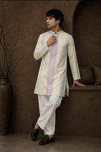A man wearing a cream-colored kurta with a vertical lavender stripe and white trousers, posing in a rustic brown room setting.