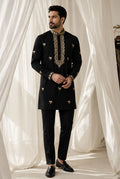 A man modeling a black kurta and trouser set with ornate gold embroidery on the collar, cuffs, and scattered motifs on the front, posed in front of draped white fabric.