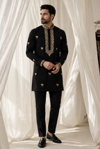 A man modeling a black kurta and trouser set with ornate gold embroidery on the collar, cuffs, and scattered motifs on the front, posed in front of draped white fabric.