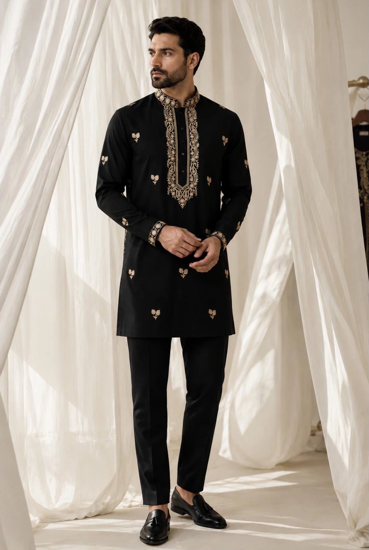 A man modeling a black kurta and trouser set with ornate gold embroidery on the collar, cuffs, and scattered motifs on the front, posed in front of draped white fabric.