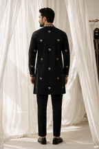 A man viewed from behind wearing a black long kurta with small gold embroidered motifs and black trousers, standing in front of draped white fabric.