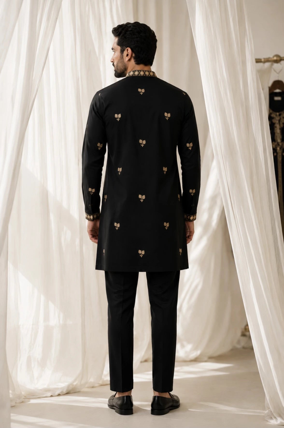A man viewed from behind wearing a black long kurta with small gold embroidered motifs and black trousers, standing in front of draped white fabric.