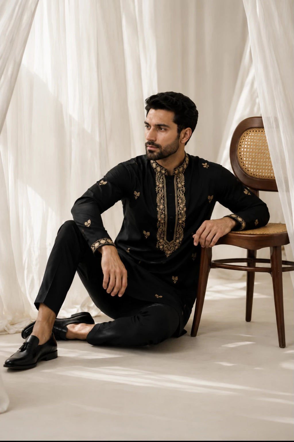 A man wearing a black traditional kurta with gold embroidery sits on a wooden chair in a softly lit room with white draped curtains.