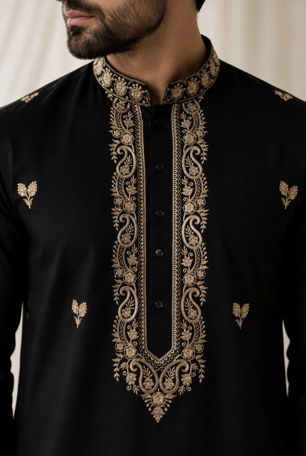 A close-up of a man wearing a black kurta with ornate gold embroidery around the mandarin collar, front placket, and small motifs on the chest.
