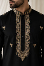 A close-up of a man wearing a black kurta with ornate gold embroidery around the mandarin collar, front placket, and small motifs on the chest.