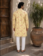 A man viewed from behind wearing a light yellow kurta with a red and green floral pattern, paired with white trousers and brown sandals, standing in an outdoor courtyard next to a large potted plant.