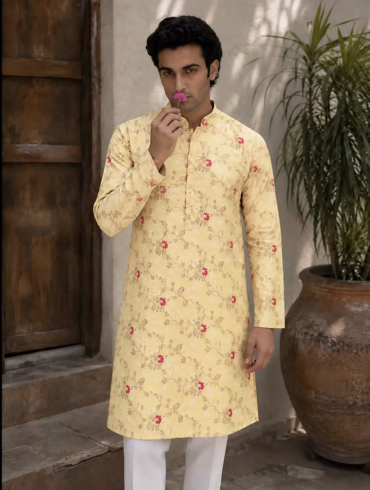 A man wearing a pale yellow long-sleeve kurta with a delicate red and beige floral pattern, paired with white trousers, standing in front of a rustic wooden door and a potted plant.