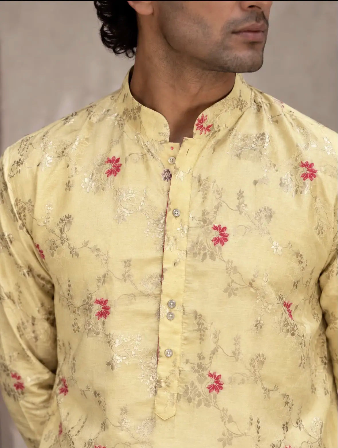 A man wearing a pale yellow kurta with a subtle metallic floral pattern and small red flower accents, shown from the chest up against a neutral studio background.