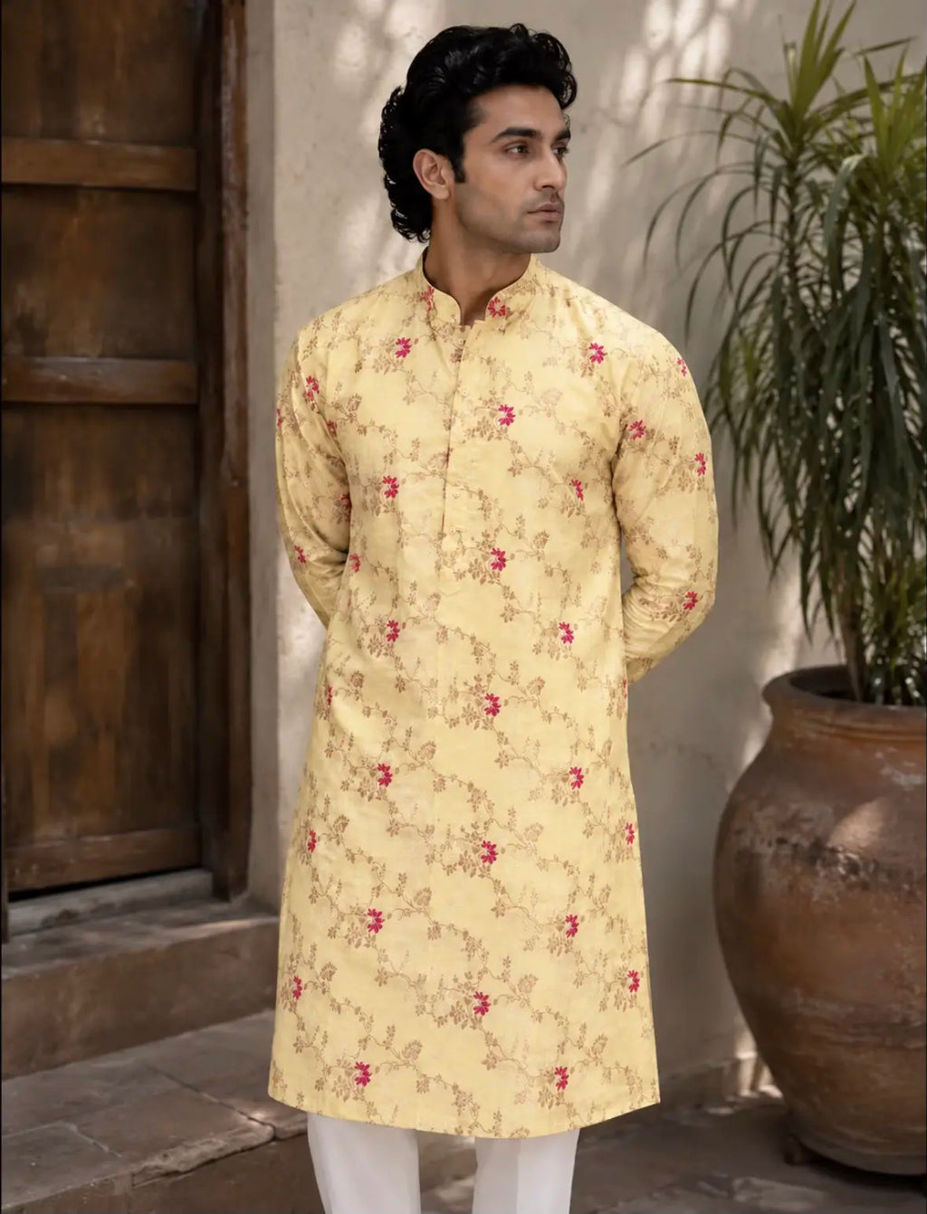 A man wearing a light yellow kurta with a red and brown floral pattern, paired with white trousers, standing in front of a rustic wooden door and a potted plant.