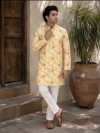 A man wearing a light yellow kurta with a red floral pattern and white trousers, standing in an outdoor courtyard next to a large potted plant.