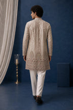 A man viewed from behind wearing a light beige embroidered sherwani jacket with white trousers, standing against a textured dark blue wall with draped white fabric and a decorative floor lamp.