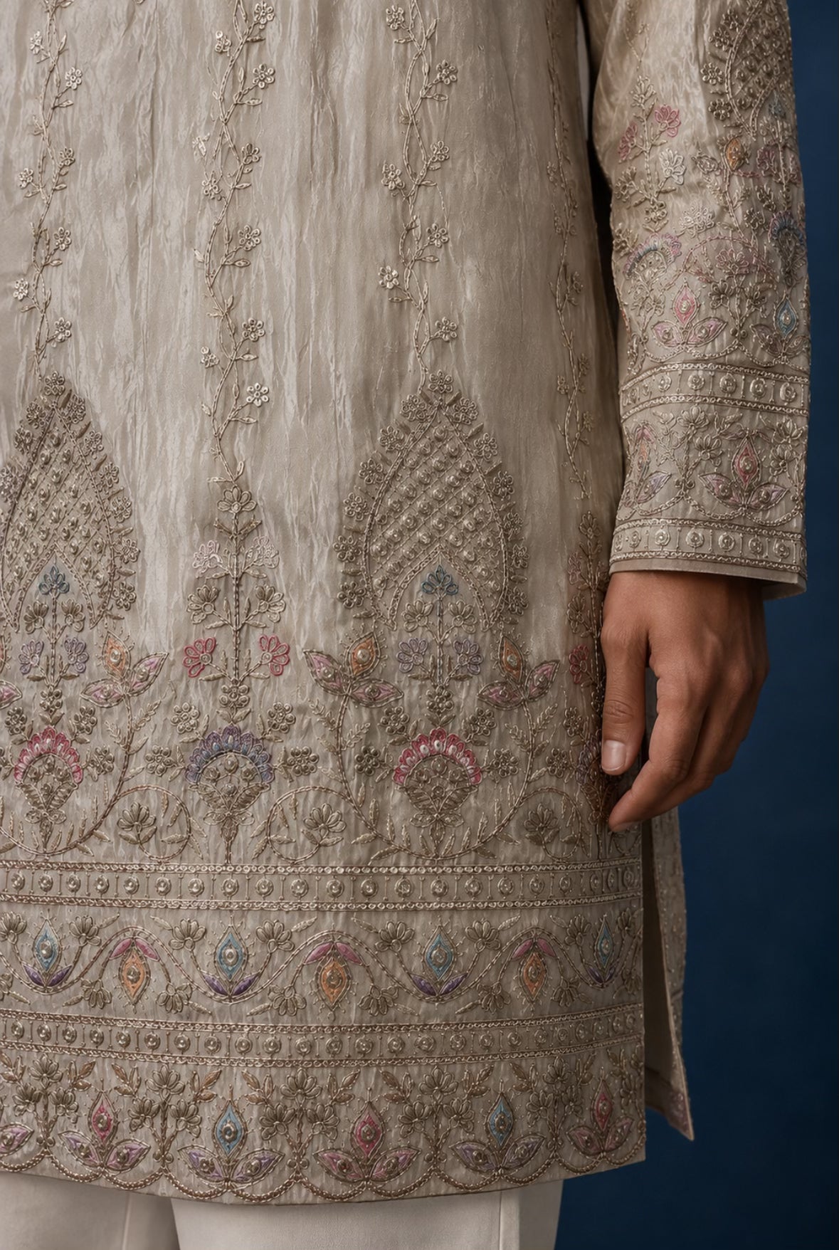 Close-up of a beige embroidered traditional kurta with intricate multicolor floral and geometric patterns, worn with white trousers against a dark blue background.