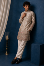 Man wearing beige embroidered kurta with arms crossed, leaning on wall