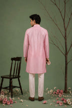 A man stands with his back to the camera wearing a light pink traditional kurta and white trousers, posed in a studio setting with a green backdrop, a dark wooden chair, a bare tree branch, and scattered pink and white flowers.