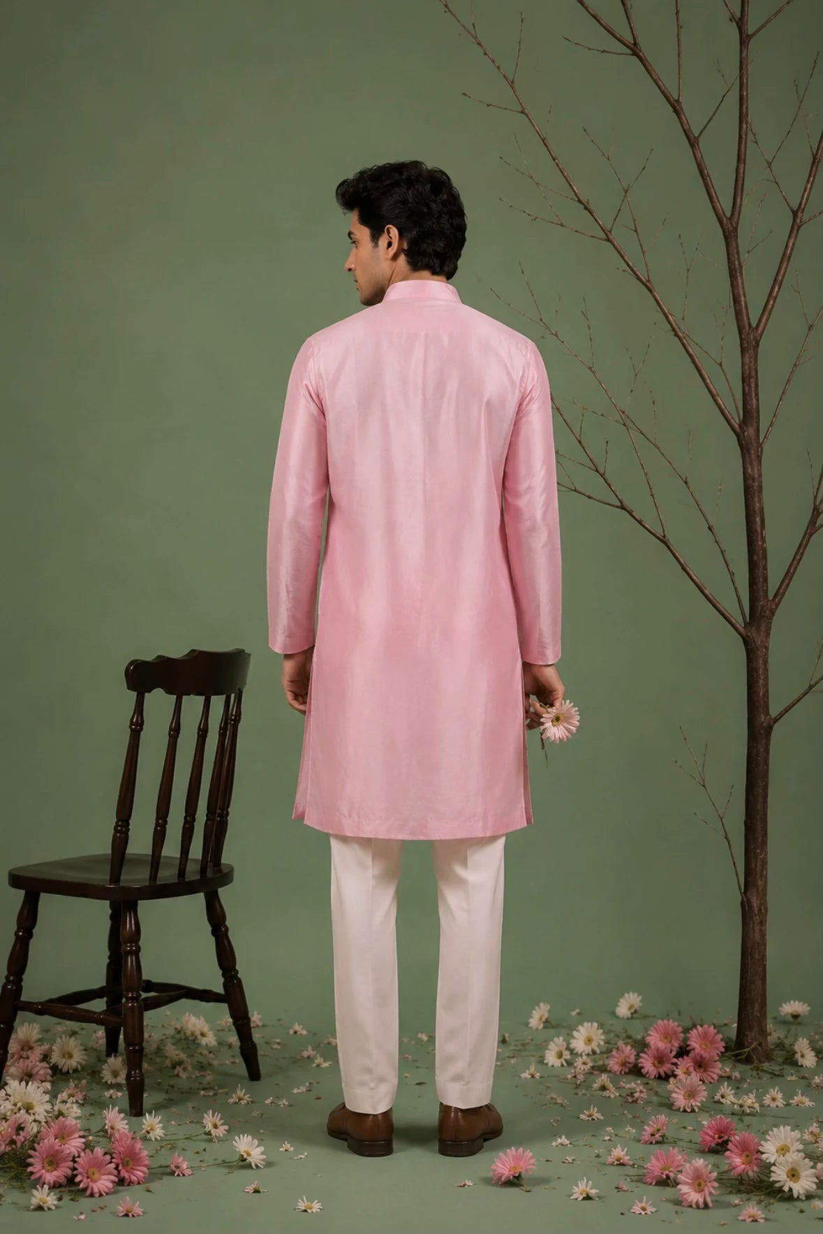 A man stands with his back to the camera wearing a light pink traditional kurta and white trousers, posed in a studio setting with a green backdrop, a dark wooden chair, a bare tree branch, and scattered pink and white flowers.