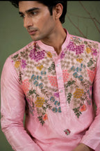 A man wearing a light pink long-sleeve kurta with colorful floral embroidery across the chest and shoulders, set against a muted olive green background.