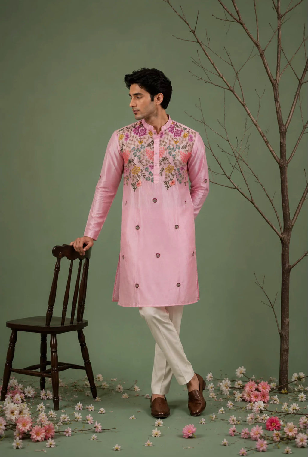 A man models a light pink traditional kurta with colorful floral embroidery on the chest and collar, paired with off-white trousers and brown shoes, standing in a studio setting with a green backdrop, a wooden chair, and scattered pink flowers.