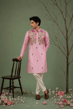 A man models a light pink traditional kurta with colorful floral embroidery on the chest and collar, paired with off-white trousers and brown shoes, standing in a studio setting with a green backdrop, a wooden chair, and scattered pink flowers.