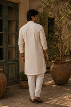 A man wearing a cream-colored kurta and matching trousers, viewed from behind as he stands in a sunlit courtyard with large terracotta pots and a rustic green door.