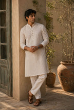 A man wearing a light beige textured kurta and white trousers stands in a sunlit courtyard next to a rustic door, with potted plants in the background.