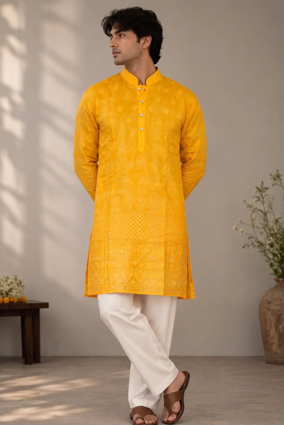 A man wearing a bright yellow embroidered kurta with white trousers and brown sandals, standing in a minimalist indoor setting with a beige wall and soft shadows.