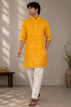 A man wearing a bright yellow kurta with white trousers and brown sandals, standing in a neutral indoor setting with soft shadows.
